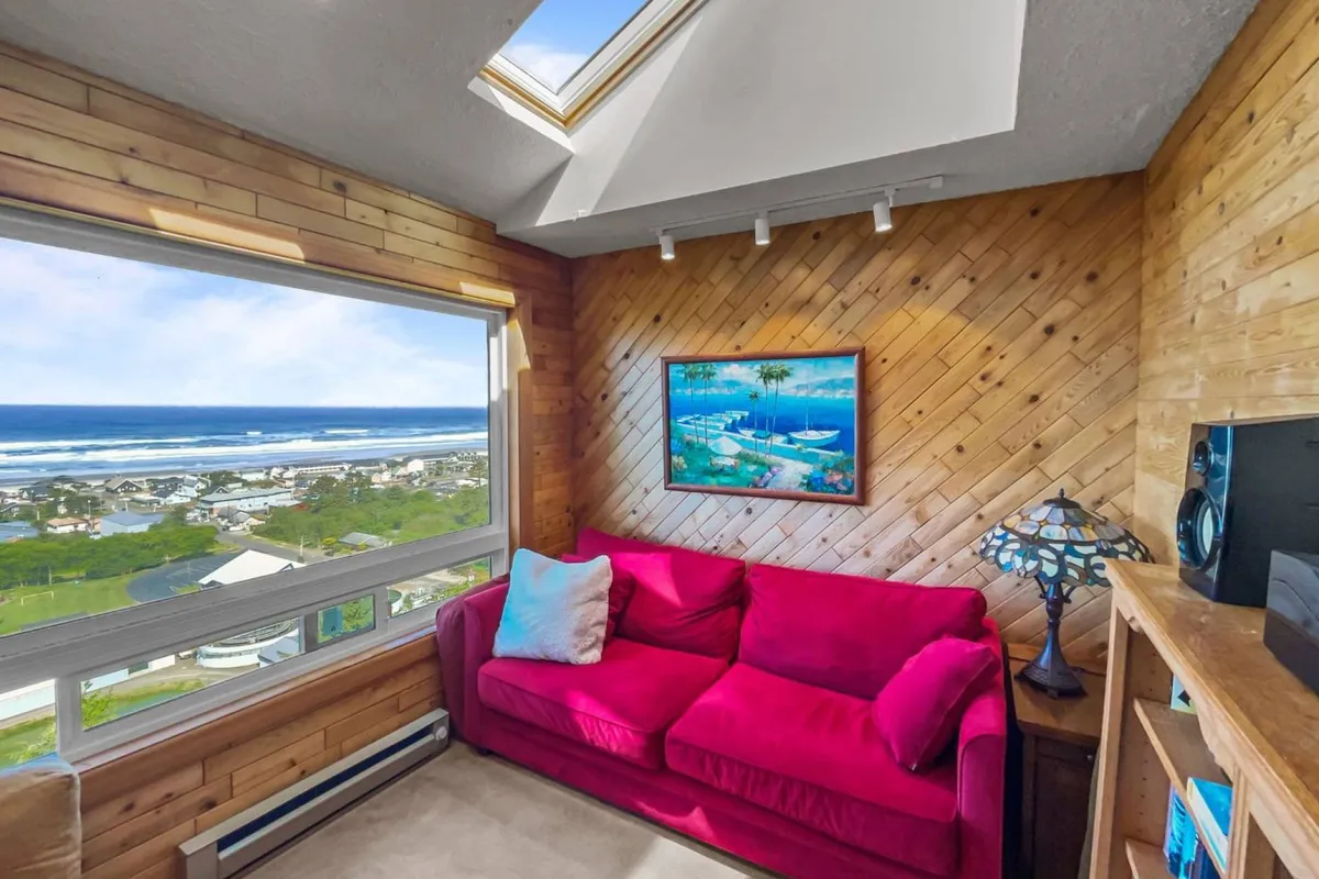 An inviting interior with a pink sofa set beside a large window framing a view of the ocean. The cozy setting is accented by a decorative painting and a table lamp, suggesting a perfect spot to unwind after outdoor adventures while still enjoying the coastal scenery.