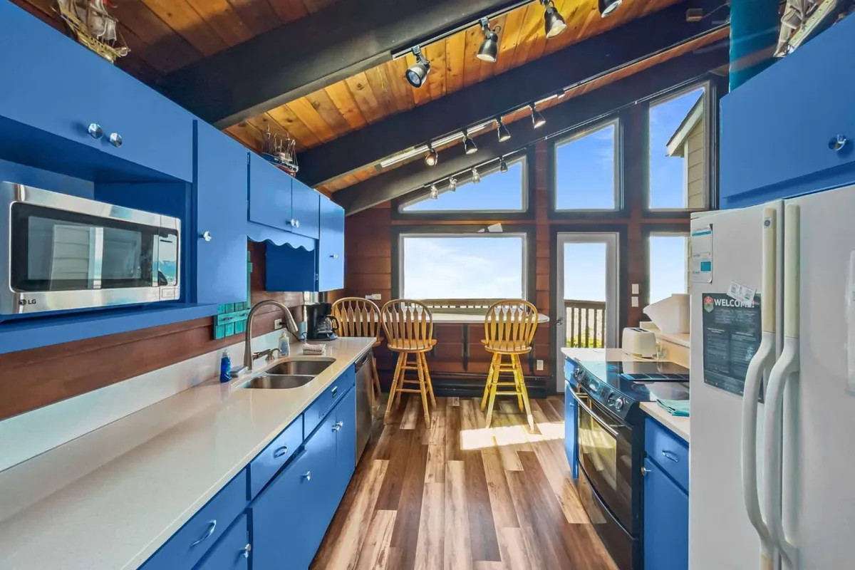 A cheerful open-plan kitchen and dining area with bright blue cabinetry, large windows offering ocean views, wooden dining chairs, and a sleek countertop. The space is filled with natural light, encouraging socializing and shared meals.