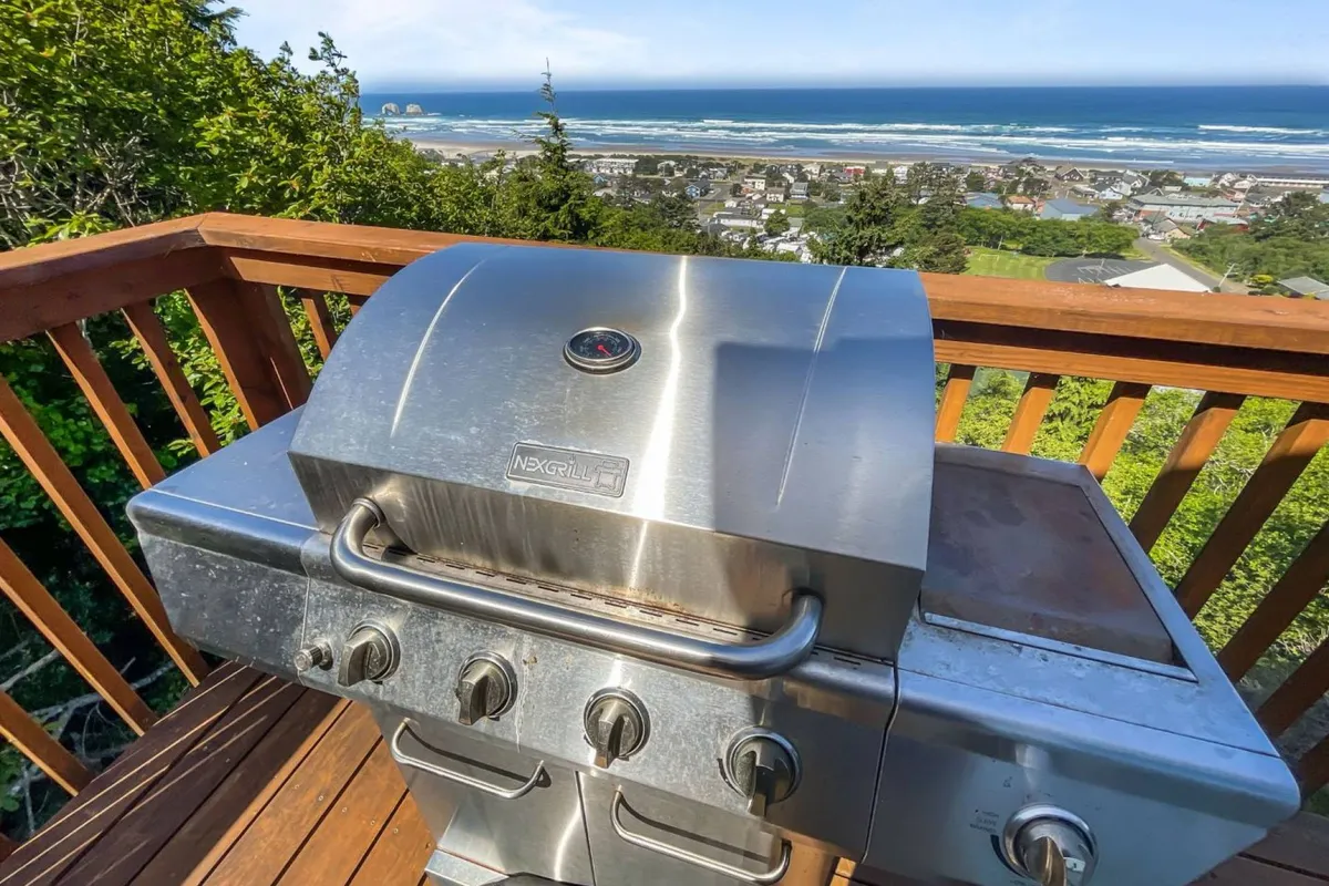 An expansive deck features a grill and outdoor dining setup, with the ocean visible in the background. The space invites social gatherings, barbecues, and memorable meals with family and friends under the open sky.
