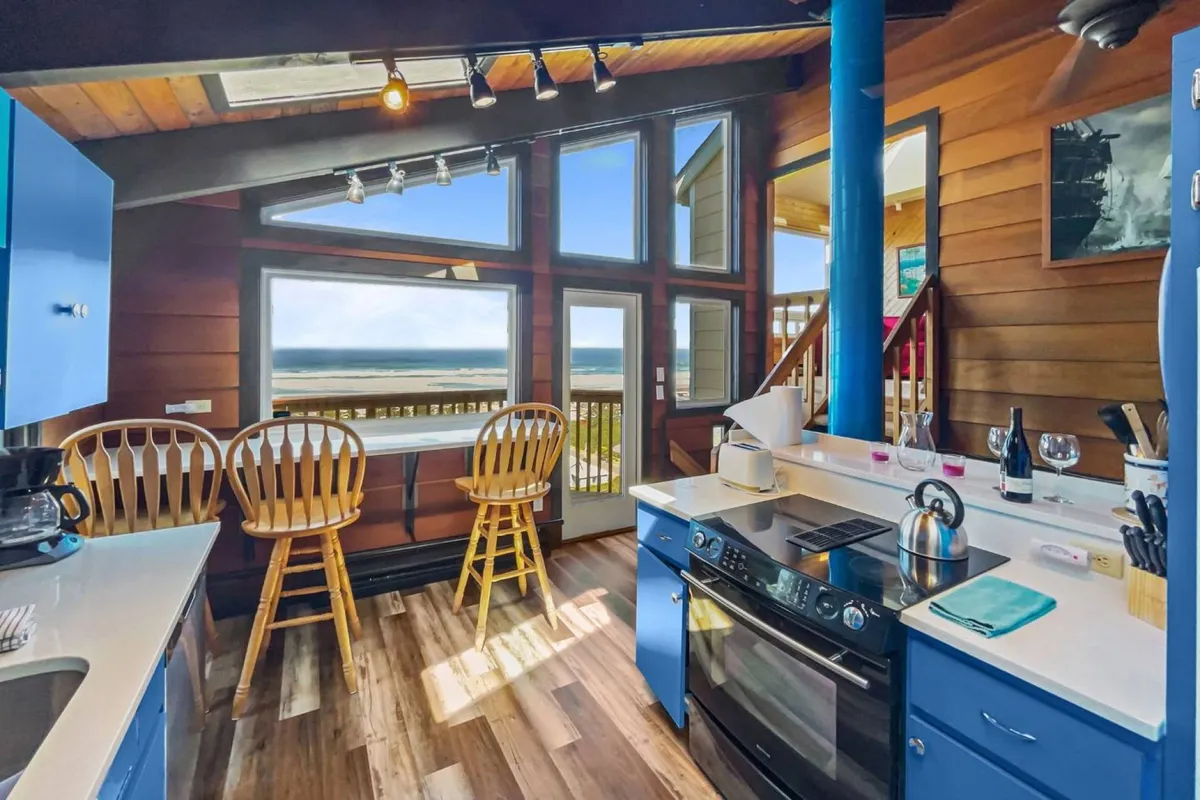 The image showcases a bright, welcoming kitchen and living space with expansive windows framing breathtaking panoramic ocean views. Blue cabinetry and wooden bar stools create a coastal-chic vibe, while natural light floods the room, inviting guests to gather, cook, and unwind as they gaze out over Rockaway Beach.