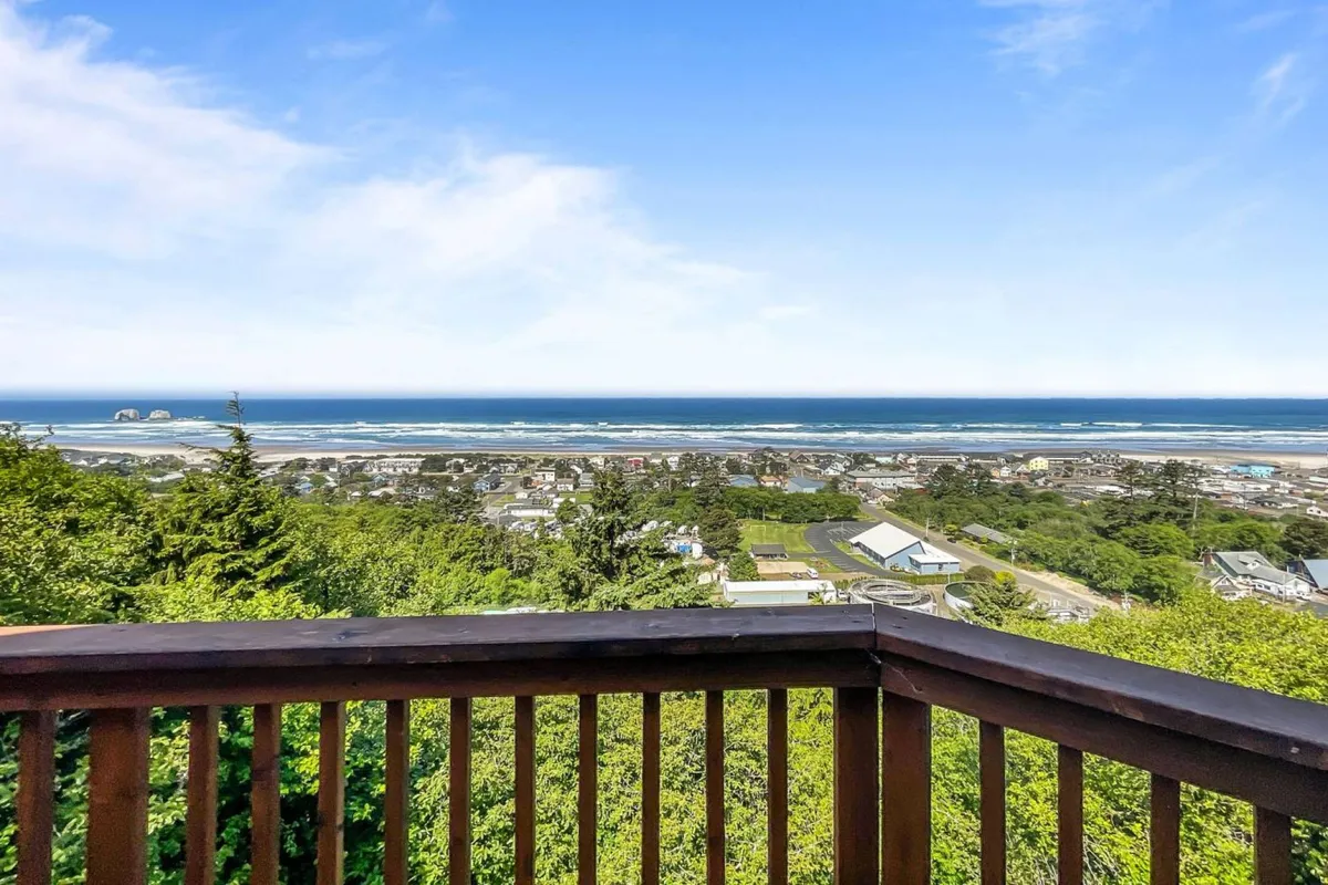 A stunning exterior view from the deck, looking out over the expansive ocean, distant coastline, and lush greenery in the foreground. The mood is tranquil, and the space is clearly suited for relaxation, meals, or stargazing under open skies.