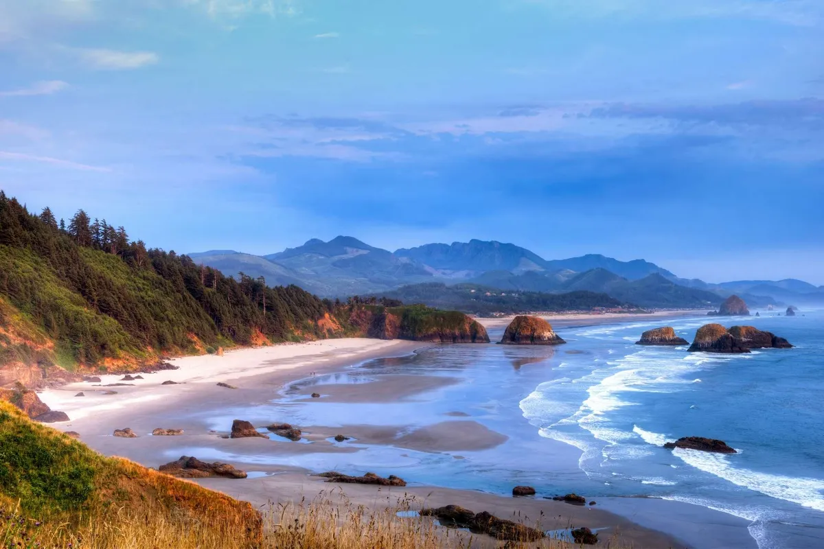 A sweeping view of a broad sandy beach bordered by the ocean, distant mountains, and rock formations. The tranquil scene captures the inviting shoreline where families and groups can enjoy activities like beachcombing, kite flying, or sunset watching.