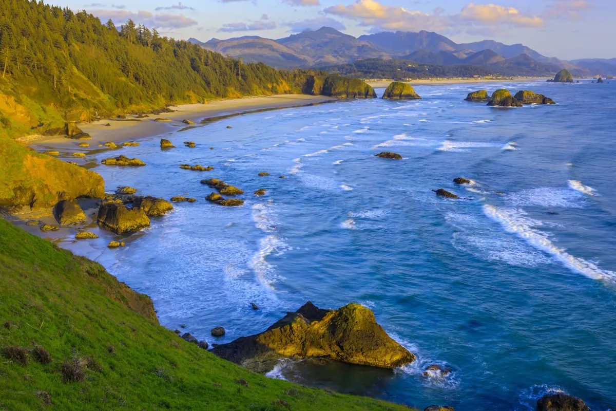 A dramatic view of the rocky coastline with waves crashing, sandy beach, and mountains in the distance. The scene is serene and inviting, suggesting opportunities for outdoor exploration and relaxation.