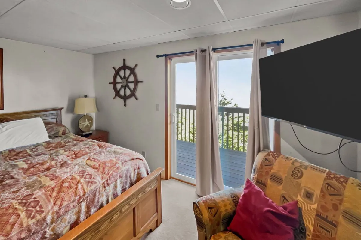 A serene bedroom featuring a bed with a patterned comforter, a window with balcony view, nautical decor including a ship's wheel, a wall-mounted TV, and a comfortable armchair. The room feels both restful and inviting, with touches that suggest a seaside retreat.