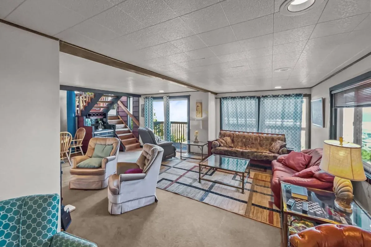 A welcoming, comfortable seating area with plush chairs and a sofa arranged near large windows that offer views to the outside. The room is bathed in natural light, and a staircase leads to the upper levels. The space feels ideal for family gatherings, relaxing, and enjoying the surrounding scenery.